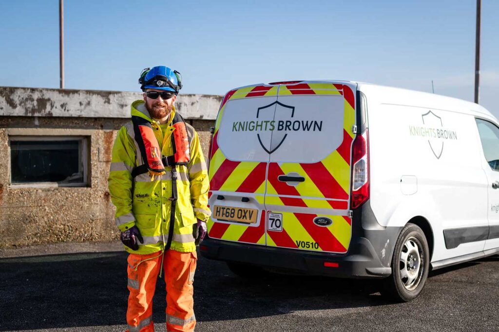 A highways technician standing infront of the back of his parked van, wearing a high vis jacket.